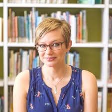 A woman with short blonde hair and glasses, wearing a sleeveless blue top, sits in front of a bookshelf filled with books.