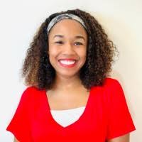 Woman with curly hair, wearing a red top and a headband, smiling in front of a plain light background.