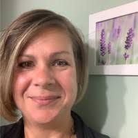 Tracy Mercier, a woman with short, light brown hair, smiles in front of a green wall with a framed picture of purple flowers.