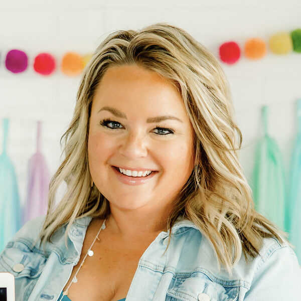A woman with blonde hair smiles at the camera, wearing a light blue denim jacket. Colorful decorations hang in the blurred background, capturing the cheerful spirit of Lucky Little Learners.